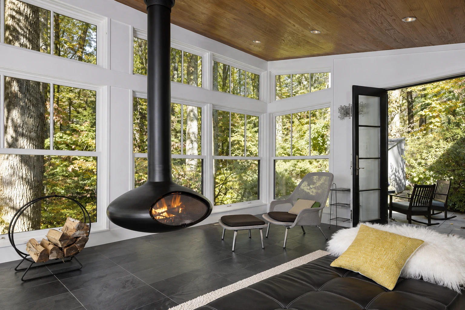 Four-season sunroom with tall windows, a suspended fireplace, and woodland views.