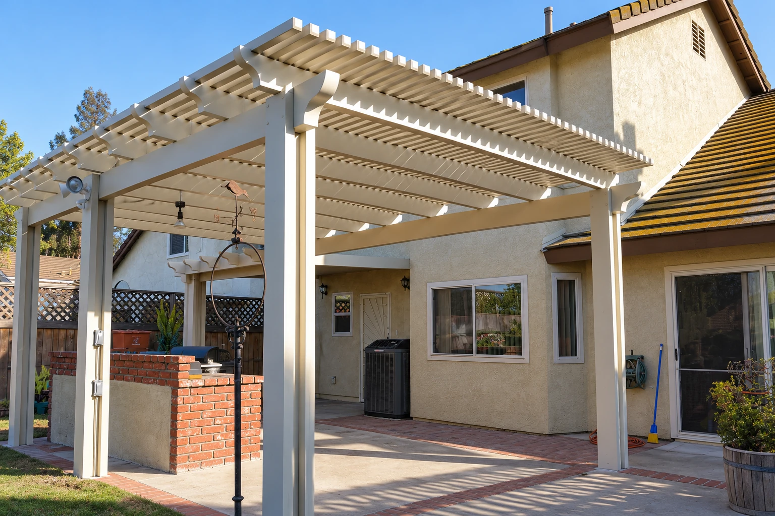 A white patio cover with slatted beams shades a paved backyard patio beside a stucco house.